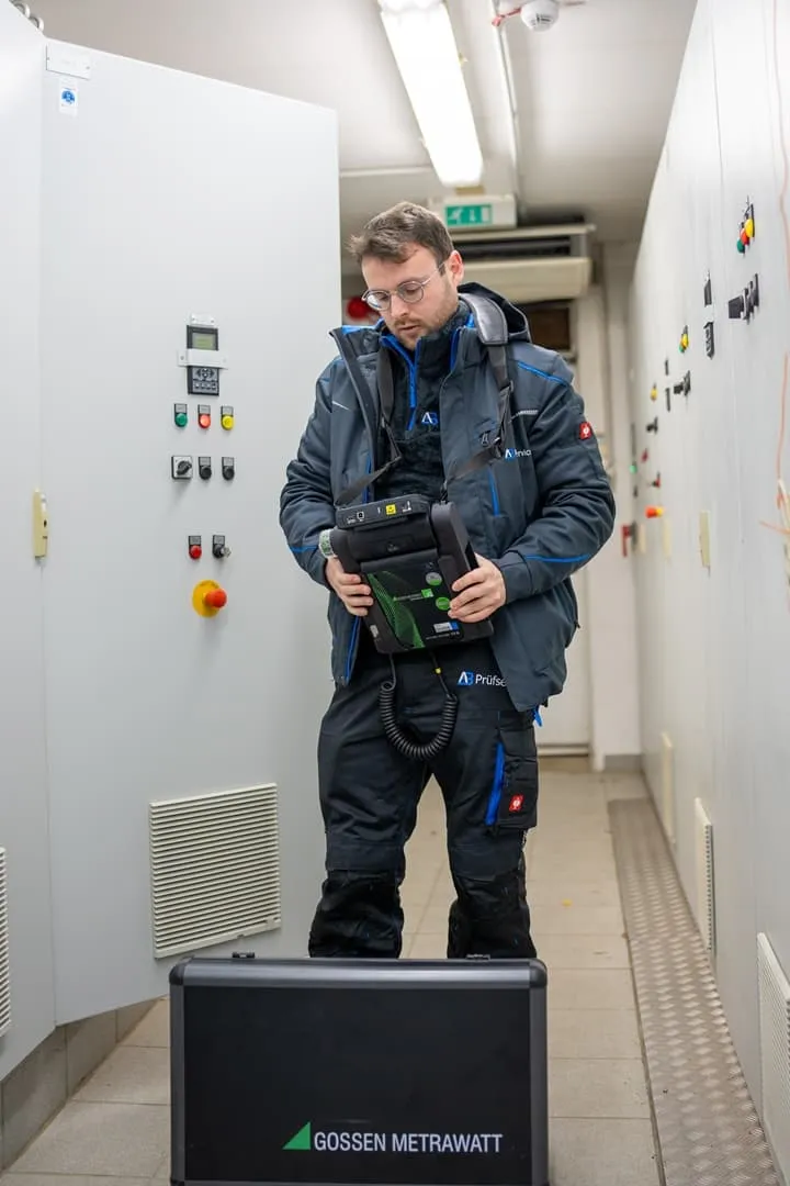 Ein Techniker in Arbeitskleidung bedient ein Handgerät in einem Kontrollraum, in dem sich Schalttafeln befinden und auf dem Boden ein Koffer von Gossen Metrawatt steht.