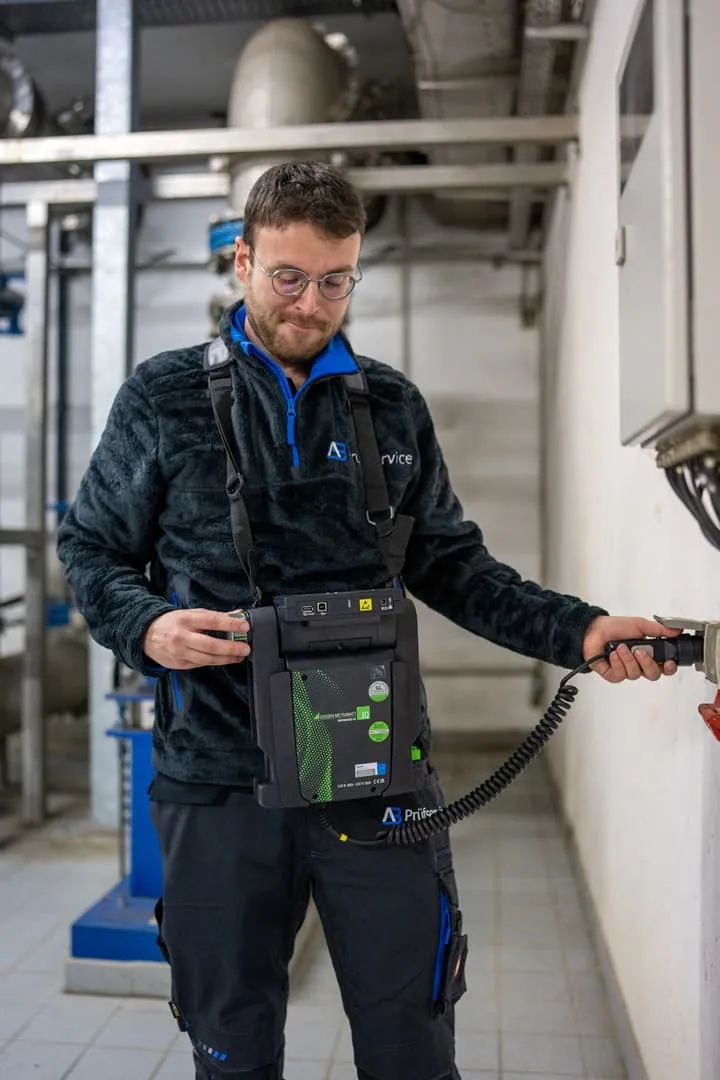 Ein Mann mit Brille und Fleecejacke bedient in einer Anlage ein tragbares elektronisches Gerät, das an Industrieanlagen angeschlossen ist.