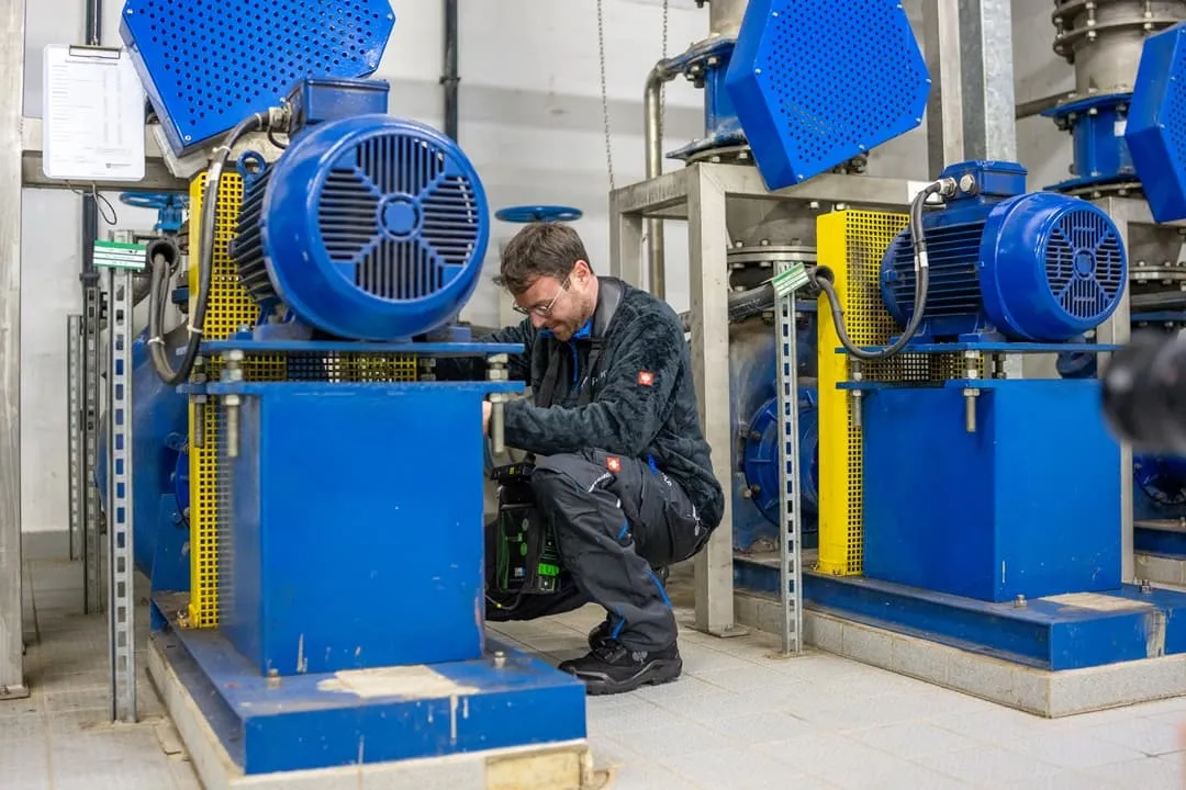 Eine Person in Arbeitskleidung überprüft oder repariert blaue Industriemaschinen in einer Halle mit Fliesenboden und sichtbaren Anlagen.