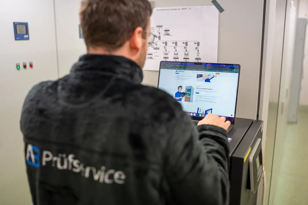 Eine Person in einer „AB Prüfservice“-Jacke arbeitet mit einem Laptop in einem Technikraum, in dem sich Schalttafeln und ein Schaltplan an der Wand befinden.