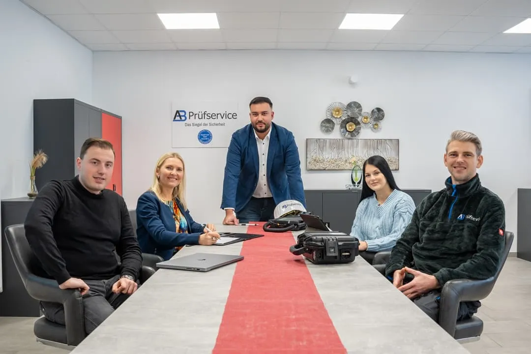 Fünf Personen sitzen und stehen um einen Konferenztisch in einem modernen Büro herum; auf dem Tisch liegen Laptops und Unterlagen, und an der Wand im Hintergrund ist ein Firmenschild mit der Aufschrift „AB Prüfservice“ zu sehen.