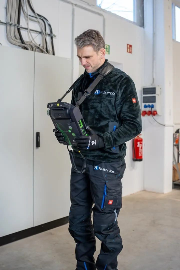 Eine Person in Arbeitskleidung bedient ein tragbares elektronisches Prüfgerät in einer industriellen Innenumgebung.