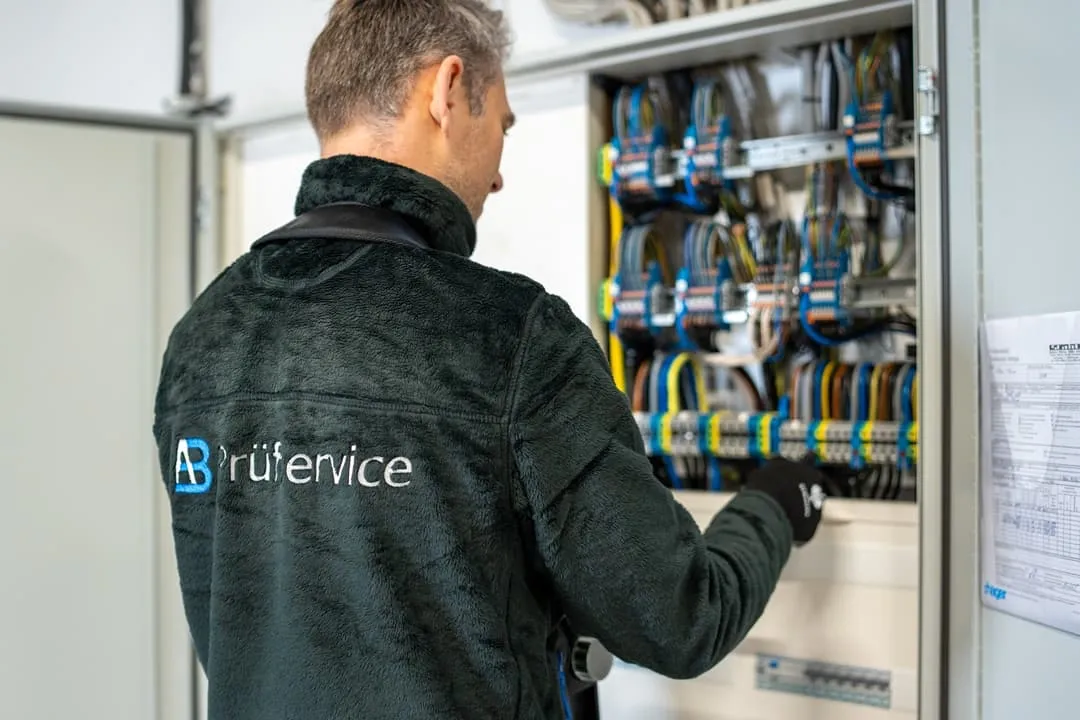 Eine Person, die eine Jacke mit der Aufschrift „AB Prüfservice“ trägt, überprüft oder arbeitet an einem offenen Schaltschrank mit verschiedenfarbigen Kabeln.