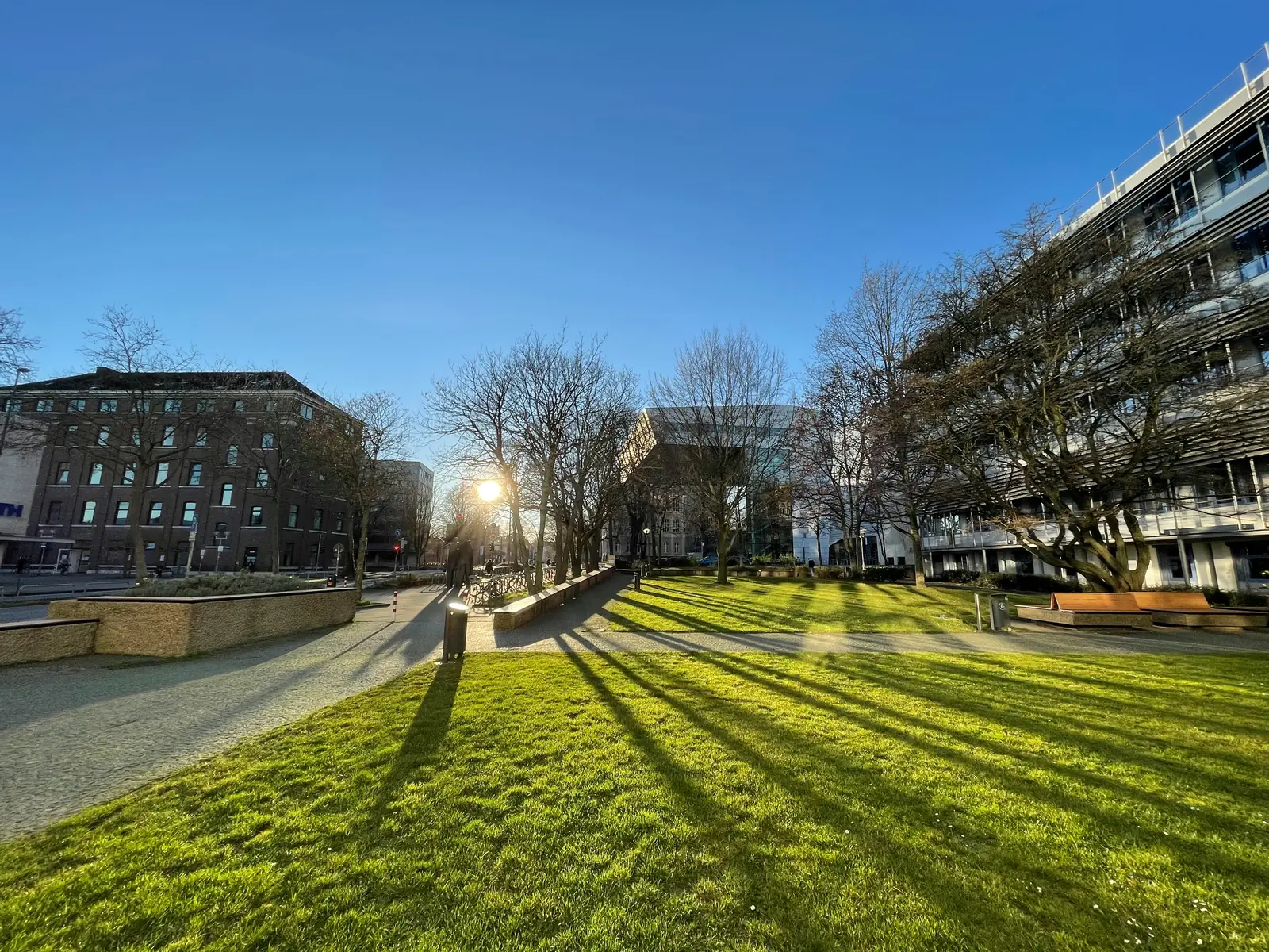 Ein Stadtpark in Aachen mit grünem Rasen, kahlen Bäumen, Bänken und umliegenden Gebäuden unter einem strahlend blauen Himmel, auf dem das Sonnenlicht lange Schatten wirft. In der Nähe sorgt die Betriebsmittelprüfung für die Sicherheit der Bevölkerung.