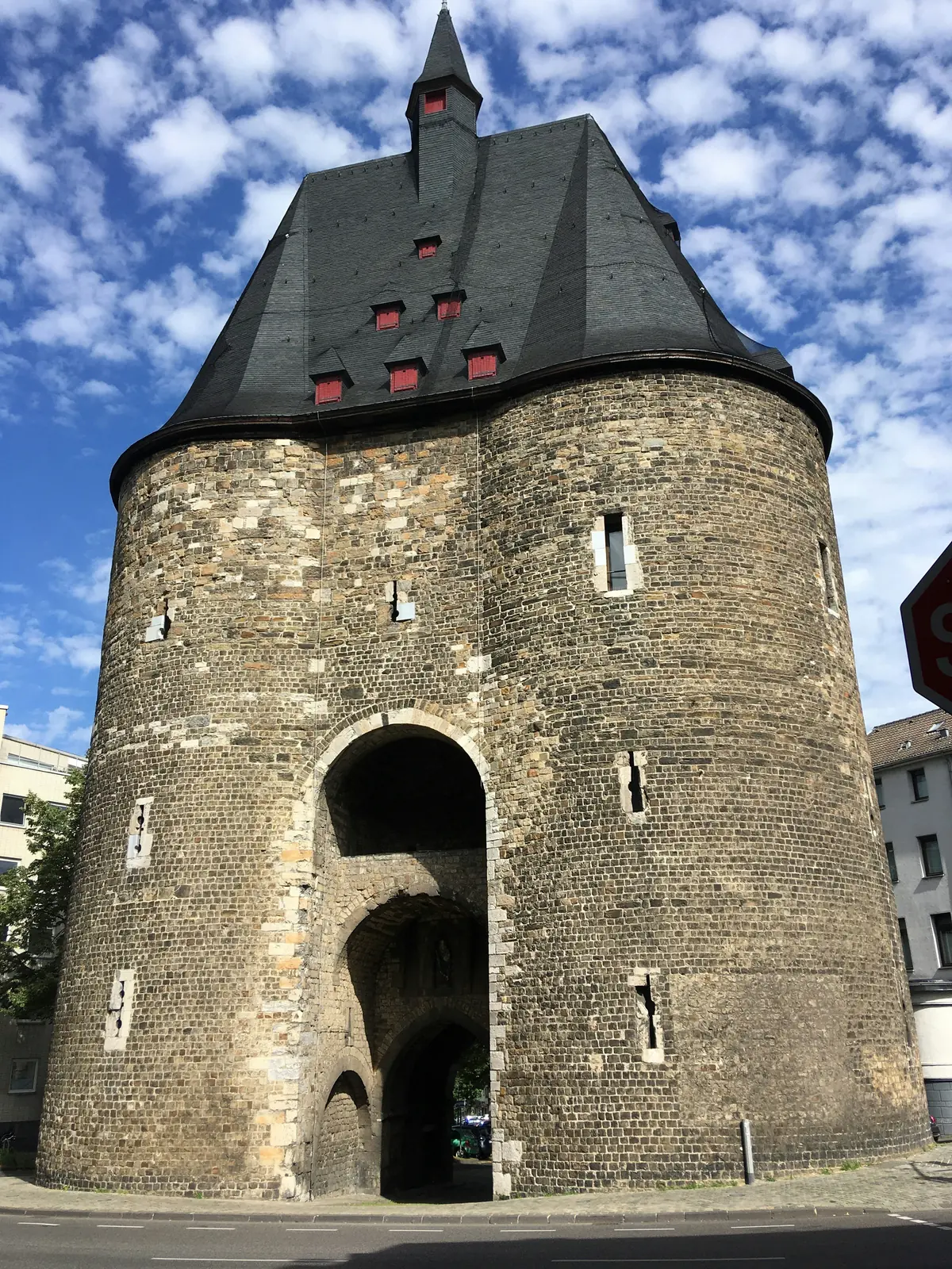 Ein historischer Stadtturm aus Stein in Aachen mit gewölbten Eingängen, kleinen, rot umrahmten Fenstern und einem steilen, dunklen Schieferdach unter einem teilweise bewölkten Himmel – eine eindrucksvolle Kulisse für die Betriebsmittelprüfung in der Region.