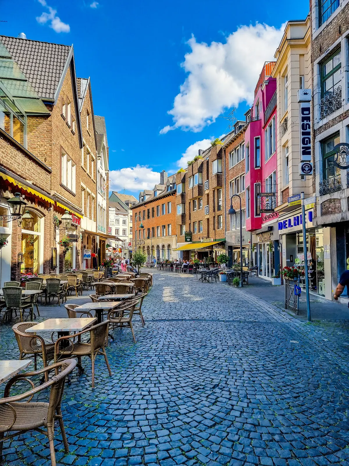 Eine Kopfsteinpflasterstraße in Aachen, gesäumt von Straßencafés und bunten Gebäuden unter einem blauen Himmel mit vereinzelten Wolken – ein Einblick in den Alltag zwischen den Terminen der Betriebsmittelprüfung.