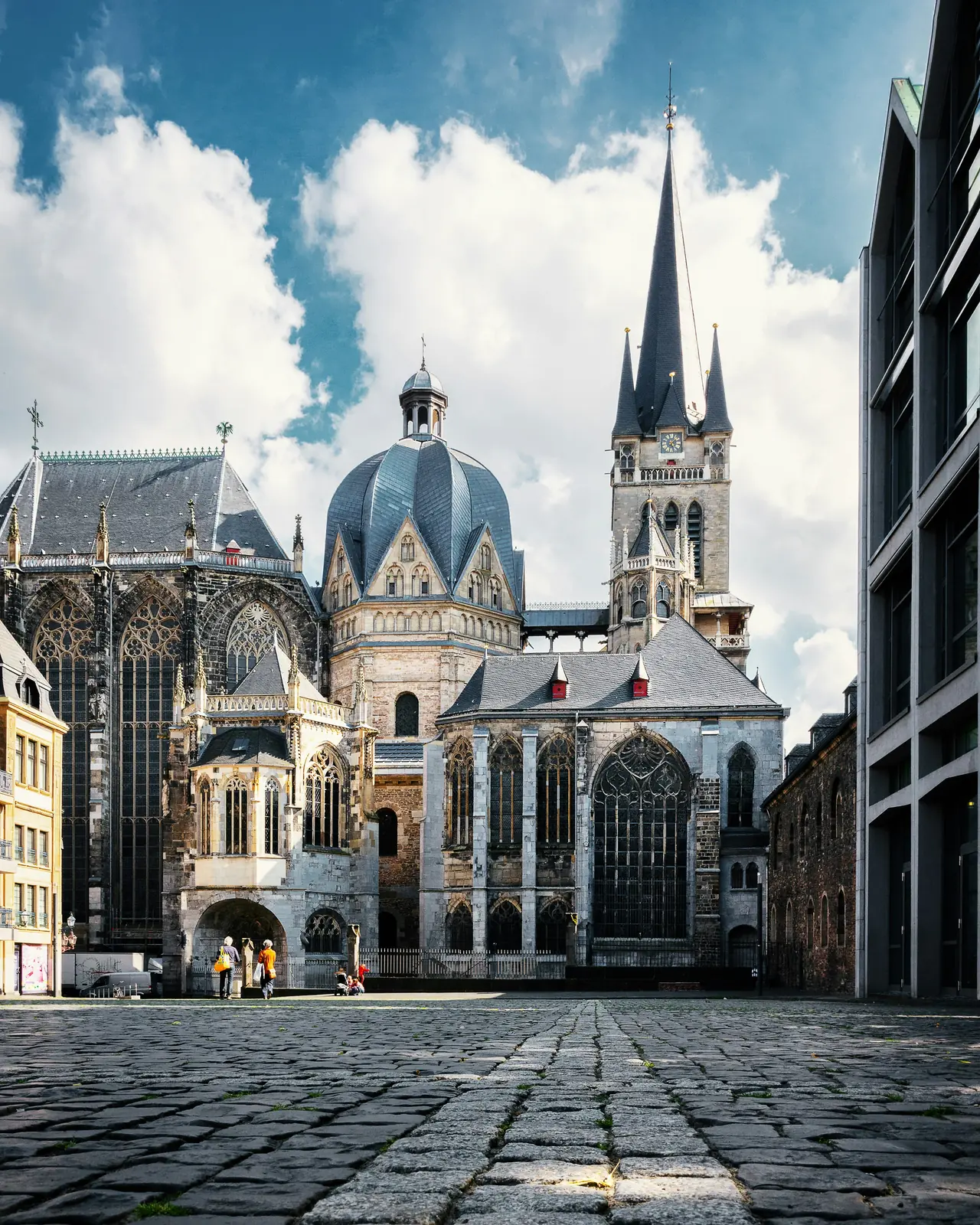 Eine historische Kathedrale mit Kuppeln und Türmen steht auf einem Kopfsteinpflasterplatz in Aachen unter einem teilweise bewölkten Himmel, während in der Nähe ein paar Menschen vorbeigehen – vielleicht unterhalten sie sich gerade über die jüngste Betriebsmittelprüfung.