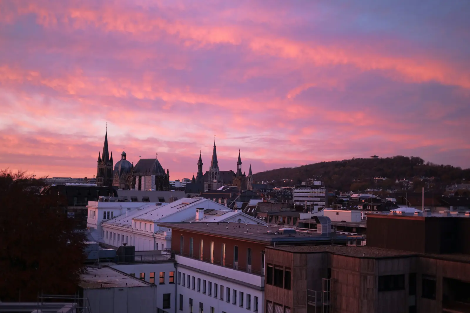 Stadtansicht von Aachen mit Kirchtürmen und Dächern vor einem rosa-orangefarbenen Sonnenuntergangshimmel und einem Hügel im Hintergrund, die den Charme der Stadt einfängt, in der Geschichte auf moderne Betriebsmittelprüfung trifft.