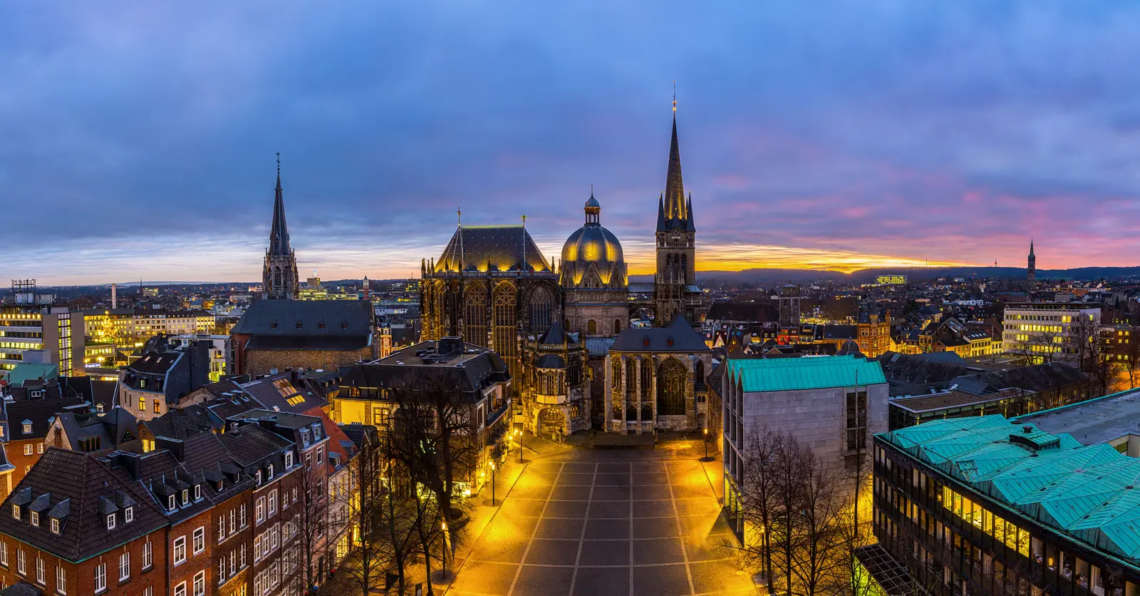 Luftaufnahme des historischen Stadtplatzes von Aachen in der Abenddämmerung, mit den markanten Türmen des Doms, beleuchteten Gebäuden und einem bewölkten Himmel mit einem Hauch von Sonnenuntergang – eine bezaubernde Szene, in der Tradition auf moderne Betriebsmittelprüfungsstandards trifft.