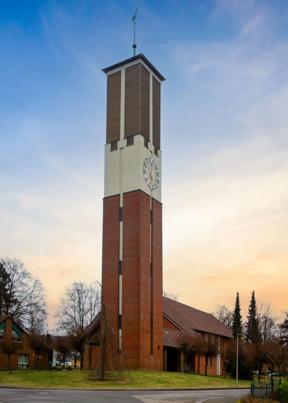 Ein hoher Backsteinbau mit weißem Uhrturm, der an ein Gebäude in Bielefeld angebaut ist, umgeben von Gras und Bäumen, unter einem bewölkten Himmel bei Sonnenuntergang – eine eindrucksvolle Kulisse für den Alltag, vielleicht sogar für wichtige Aufgaben wie die Betriebsmittelprüfung.