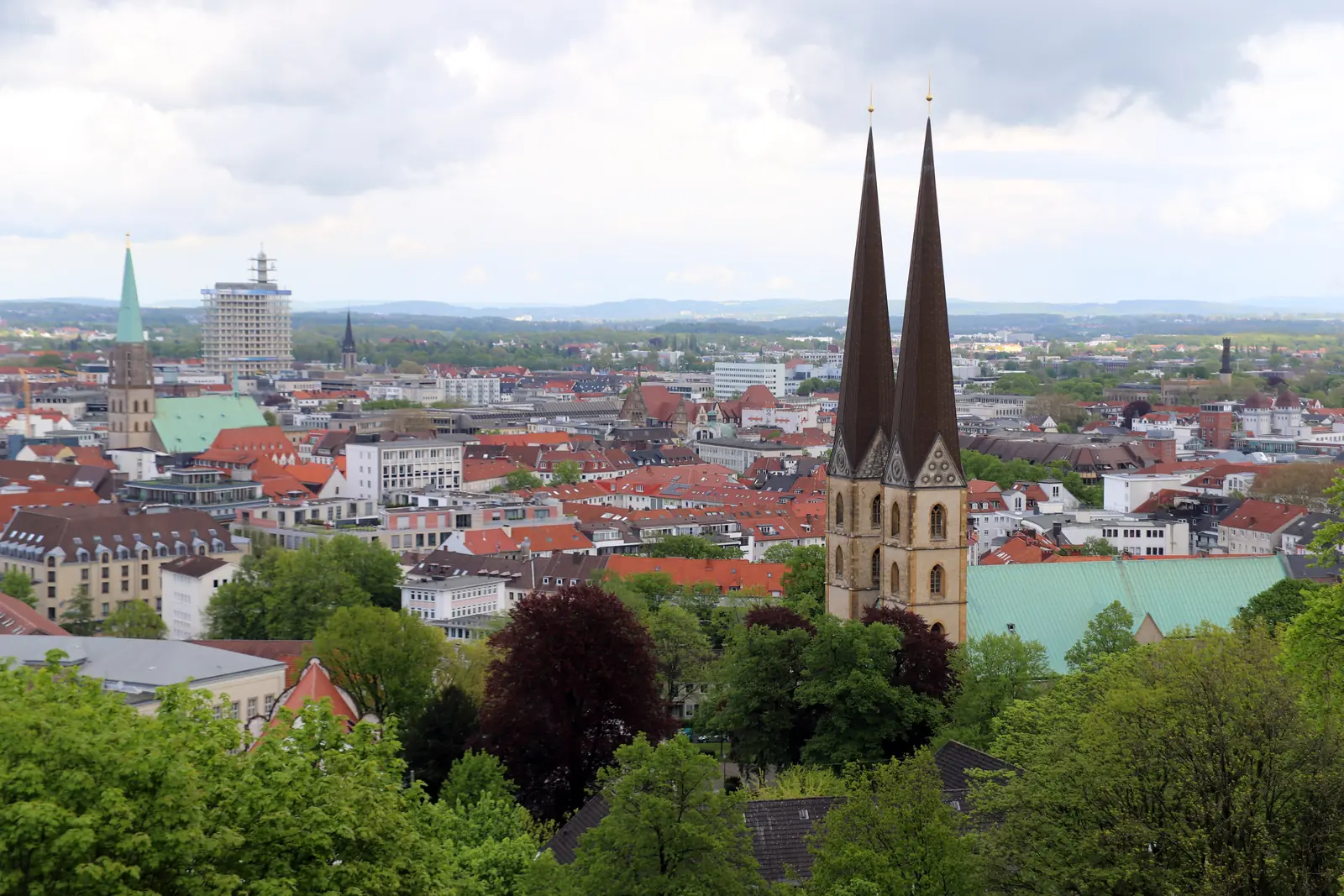 Ein Stadtbild mit einer Kirche mit zwei Türmen in Bielefeld, Gebäuden mit roten Dächern und Grünflächen im Vordergrund unter einem bewölkten Himmel – eine harmonische Mischung aus historischer Architektur und modernem Leben, in der sogar die Betriebsmittelprüfung für einen reibungslosen Ablauf in der Stadt sorgt.