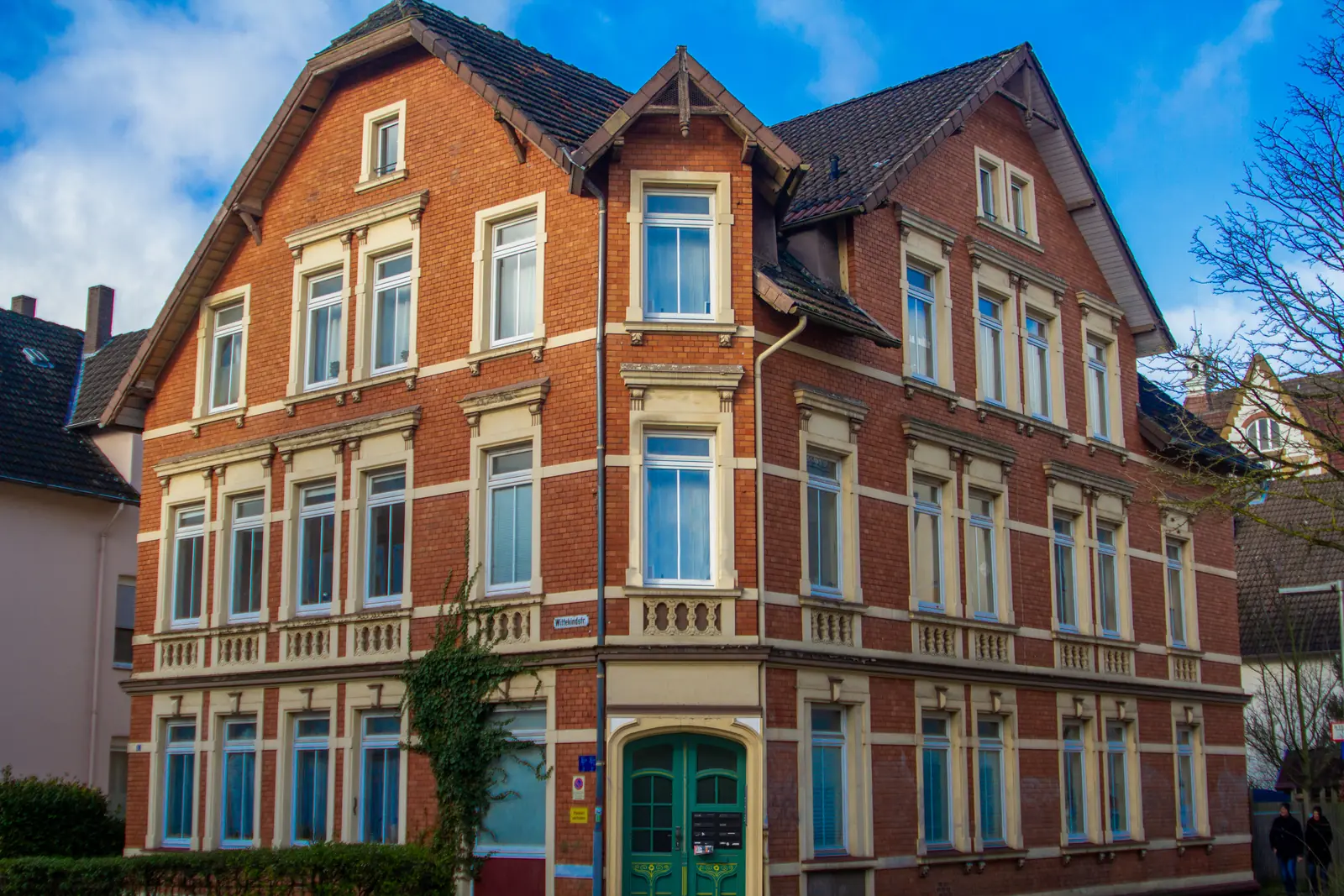 Ein dreistöckiges Backsteingebäude mit weißen Zierleisten, mehreren großen Fenstern, einer grünen Doppeltür als Eingang und einem schrägen, dunklen Dach unter einem teilweise bewölkten Himmel in Bielefeld.