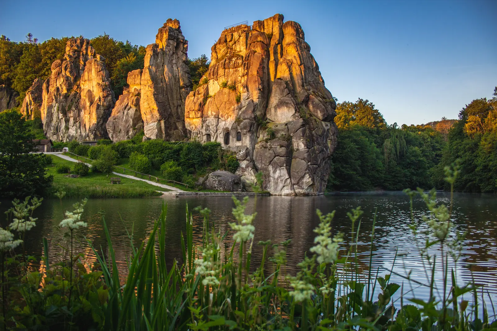 Hohe Felsformationen ragen neben einem ruhigen See empor, umgeben von grüner Vegetation und Bäumen unter einem klaren Himmel bei Sonnenuntergang – ein Bild, das die friedliche Schönheit in der Nähe von Bielefeld einfängt.