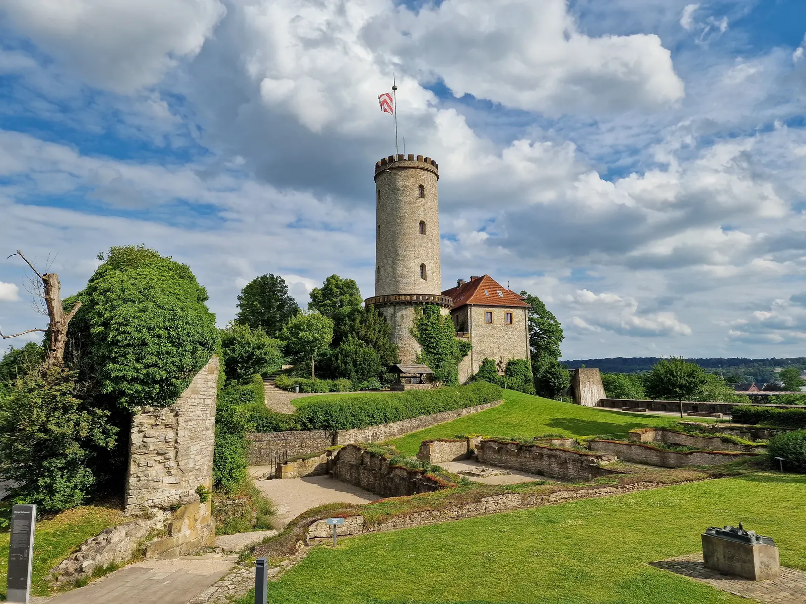 Neben einem kleinen Gebäude steht ein runder Steinturm mit einer Fahne, umgeben von Steinmauern und grünen Rasenflächen unter dem teilweise bewölkten Himmel über Bielefeld.