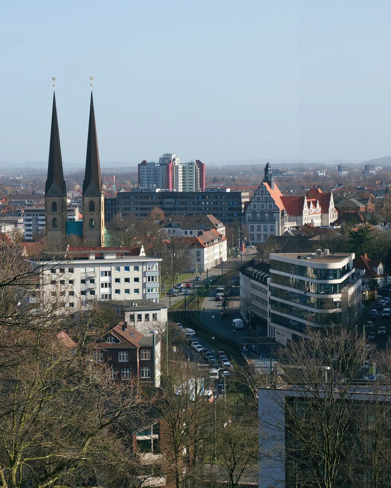 Stadtansicht von Bielefeld mit zwei Kirchtürmen, die sich vor dem Hintergrund moderner und historischer Gebäude, kahlen Bäumen und einem klaren Himmel erheben – ein Symbol für die harmonische Verbindung von Tradition und Fortschritt, das an eine Betriebsmittelprüfung erinnert, bei der Sicherheit und Innovation im Gleichgewicht stehen.