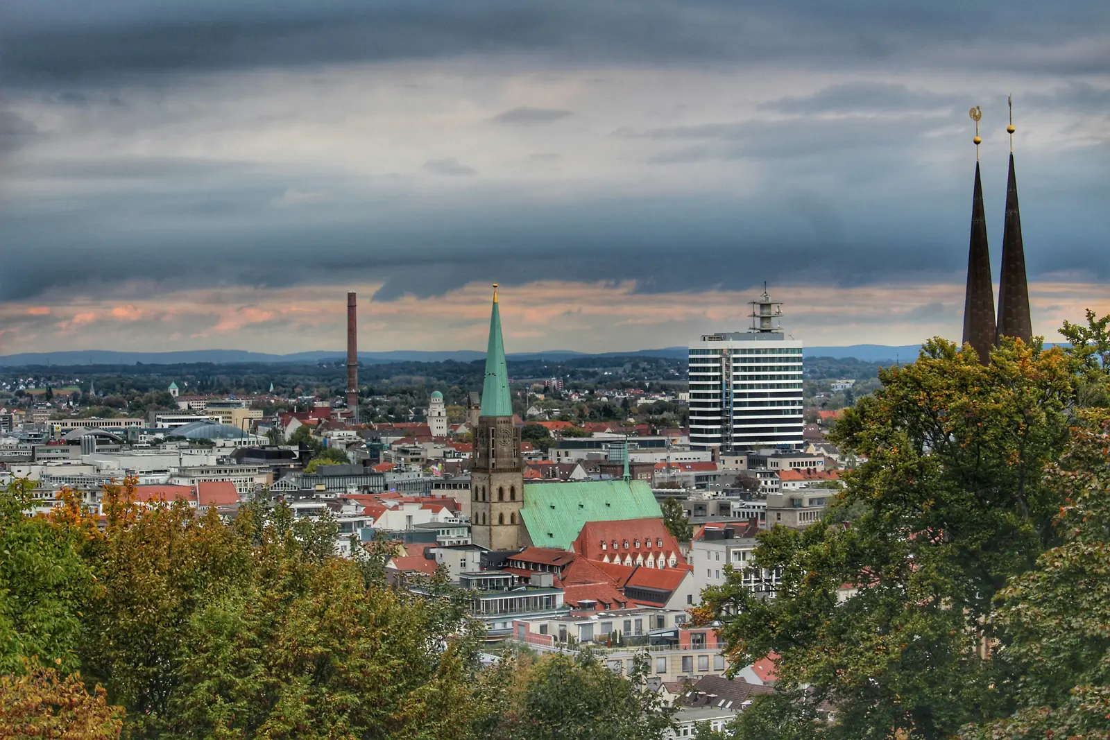 Stadtansicht von Bielefeld mit einer markanten Kirche mit begrüntem Dach, modernen Gebäuden und zwei hohen Türmen, die sich vor einem bewölkten Himmel erheben, eingerahmt von Bäumen im Vordergrund – eine Szene, die auf subtile Weise den Fleiß der Stadt und ihre Liebe zum Detail, wie beispielsweise die Betriebsmittelprüfung, andeutet.