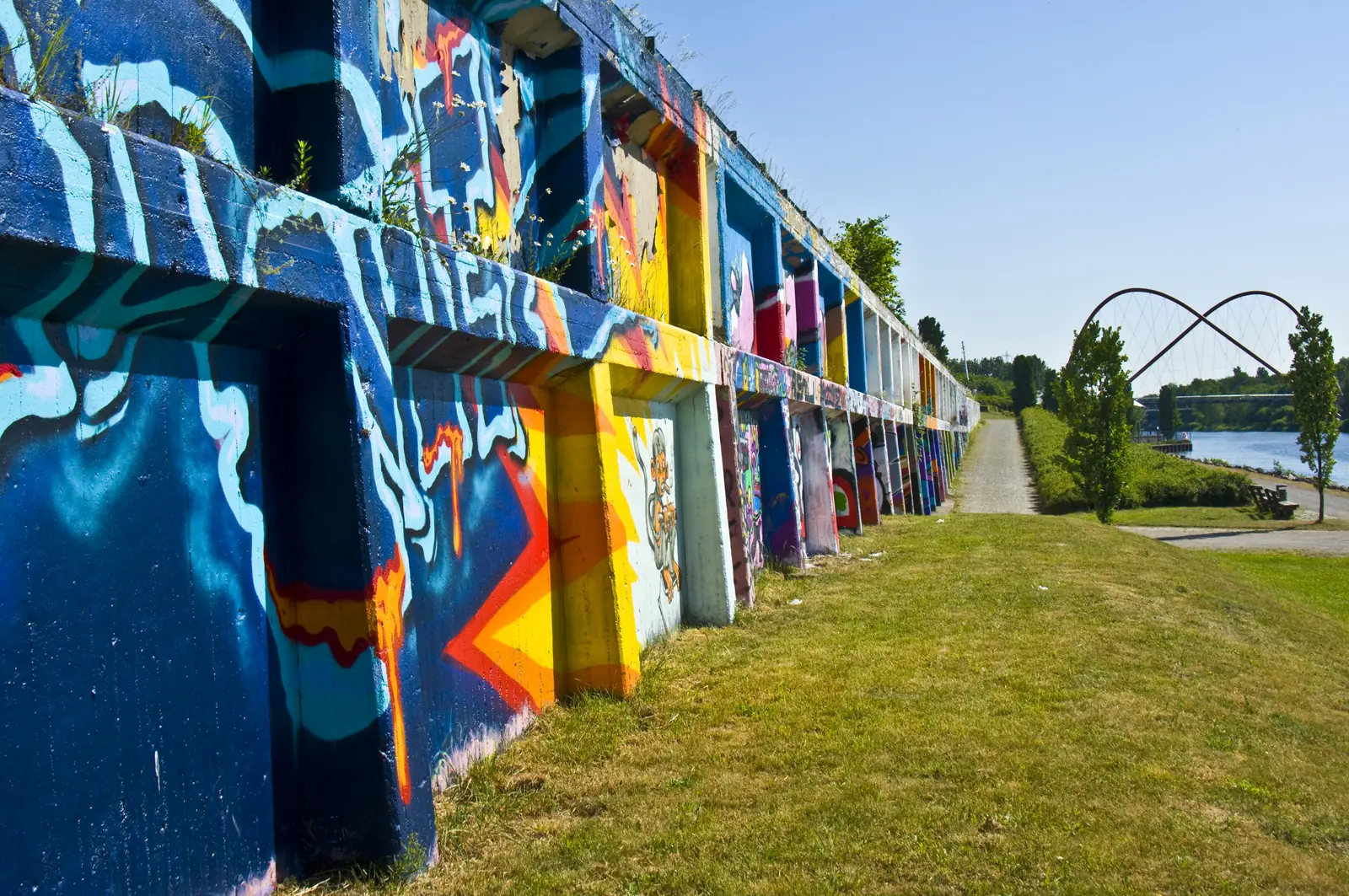 Eine mit bunten Graffiti verzierte Betonmauer neben einem grasbewachsenen Weg in Gelsenkirchen, im Hintergrund unter strahlend blauem Himmel sind ein Fluss, Bäume und eine moderne Bogenbrücke zu sehen – eine lebendige Stadtszene, in der Kunst und Industrie inmitten der routinemäßigen Betriebsmittelprüfung aufeinandertreffen.