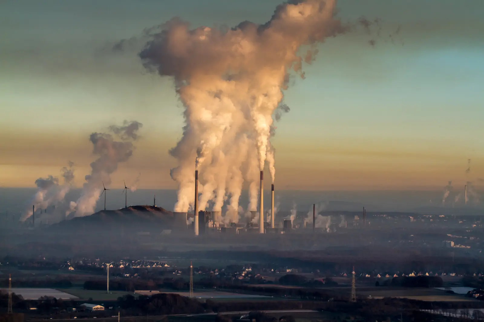 Rauch und Dampf steigen aus zahlreichen Industrieschornsteinen in Gelsenkirchen auf, im Hintergrund sind Windkraftanlagen zu sehen und im Vordergrund ein Wohngebiet, was den Kontrast zwischen traditioneller Industrie und moderner Energie verdeutlicht – während eine gründliche Betriebsmittelprüfung die Betriebssicherheit gewährleistet.