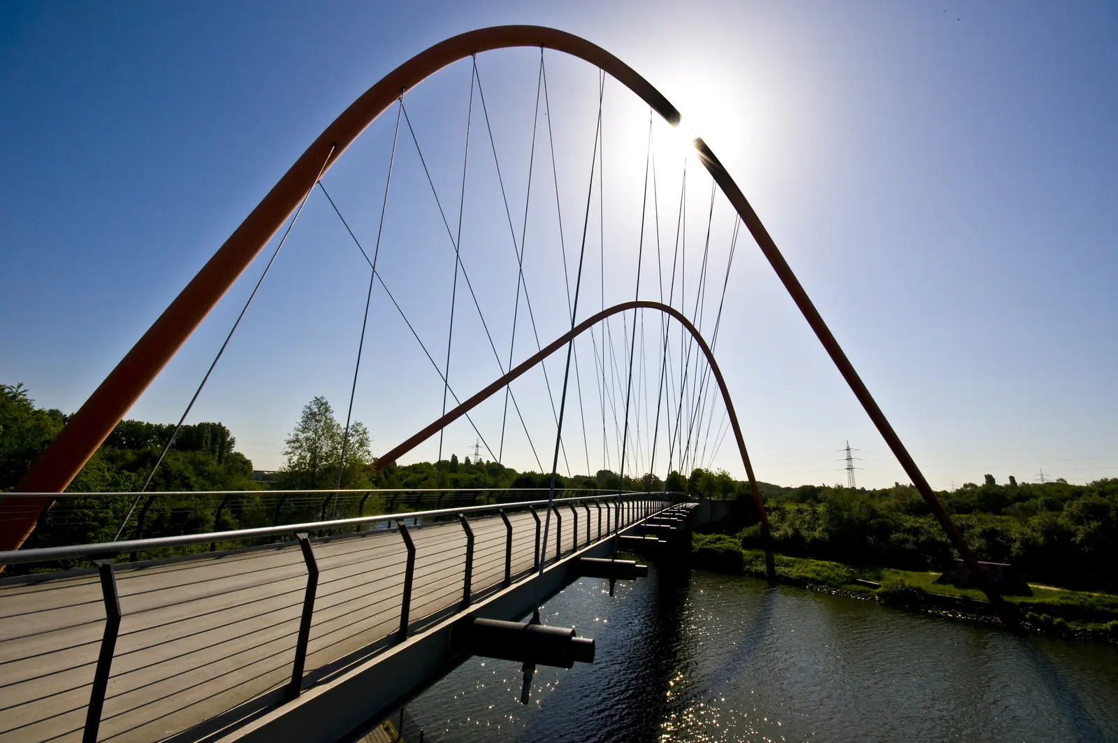 Eine moderne Fußgängerbrücke mit orangefarbenen Bögen und Seilen überspannt einen Fluss in Gelsenkirchen, während hinter dem Bauwerk Sonnenlicht scheint und im Hintergrund Grünflächen zu sehen sind.