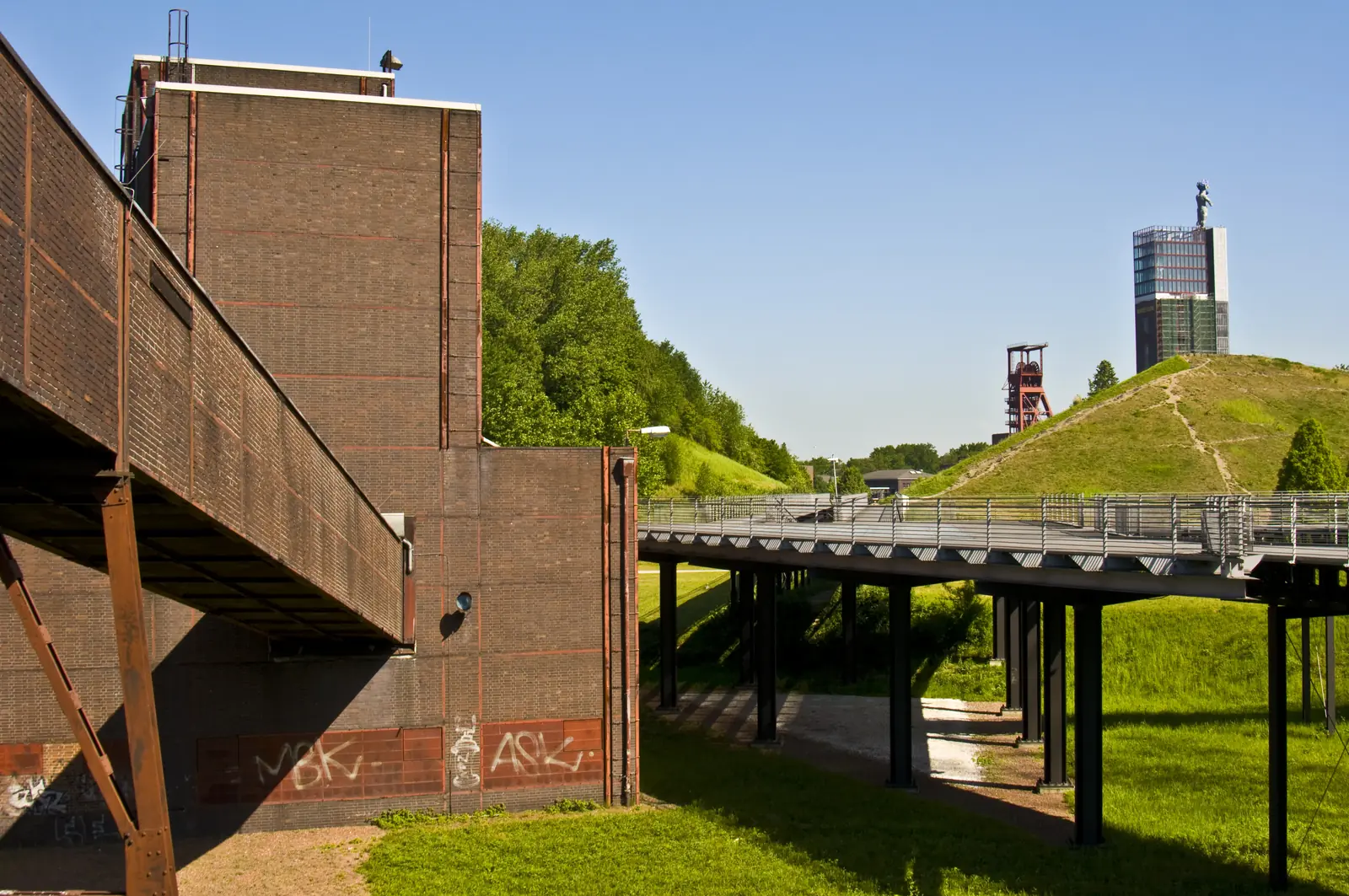 Ein modernes Industriegebäude in Gelsenkirchen mit Graffiti an den Wänden steht neben einem erhöhten Fußweg, im Hintergrund sind unter klarem Himmel grasbewachsene Hügel und ein weiteres Industriegebäude zu sehen – möglicherweise für die Betriebsmittelprüfung genutzt.