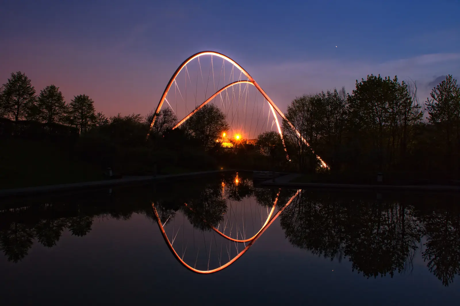 Eine moderne Bogenbrücke in Gelsenkirchen wird in der Abenddämmerung beleuchtet und spiegelt sich in einem ruhigen Fluss wider, während sich die Silhouetten der Bäume vor dem Himmel bei Sonnenuntergang abzeichnen.