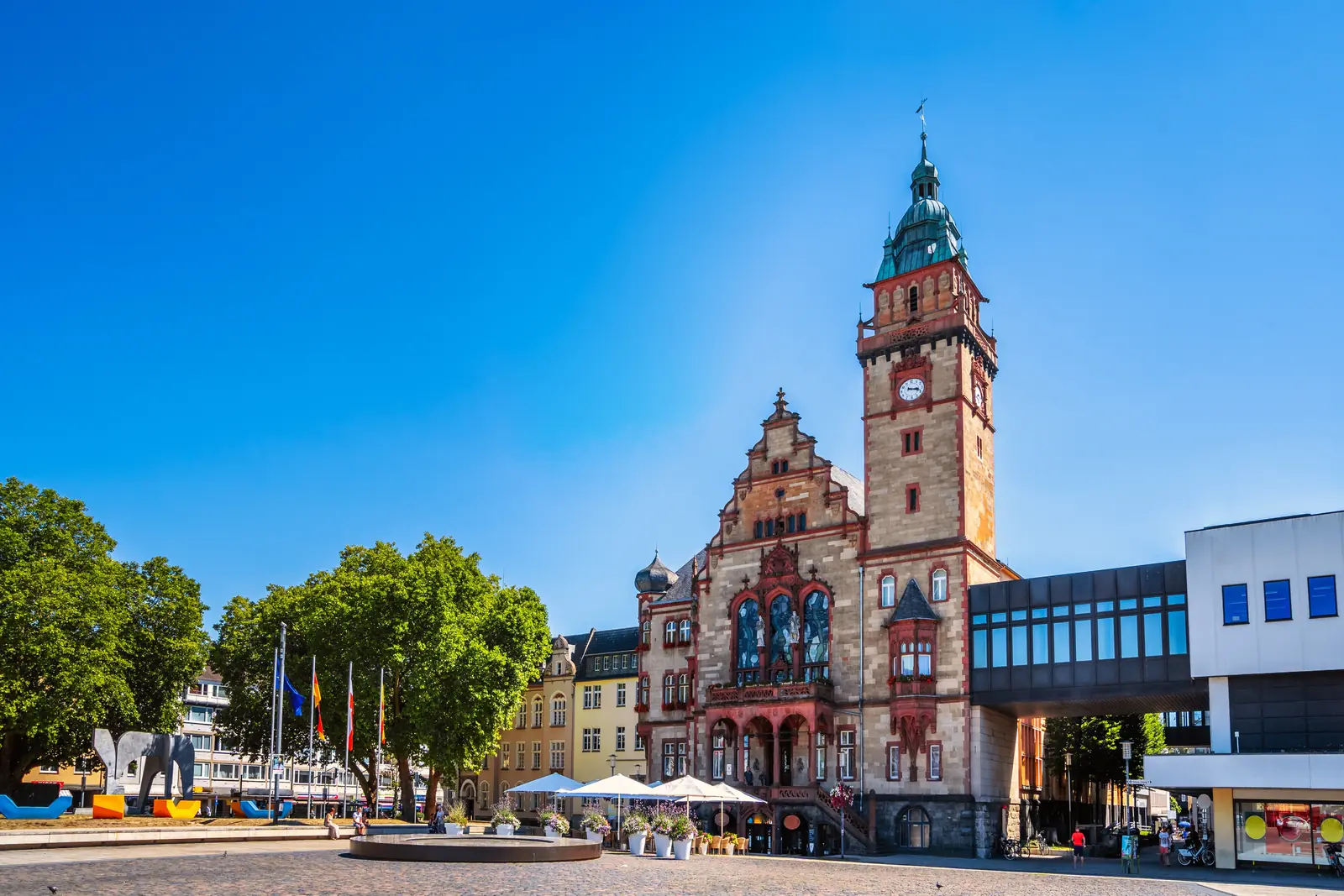 Auf einem sonnigen öffentlichen Platz in Mönchengladbach steht ein historisches Rathaus mit einem Uhrturm und Details aus rotem Backstein, geschmückt mit Fahnen, Bäumen und Sitzgelegenheiten im Freien.