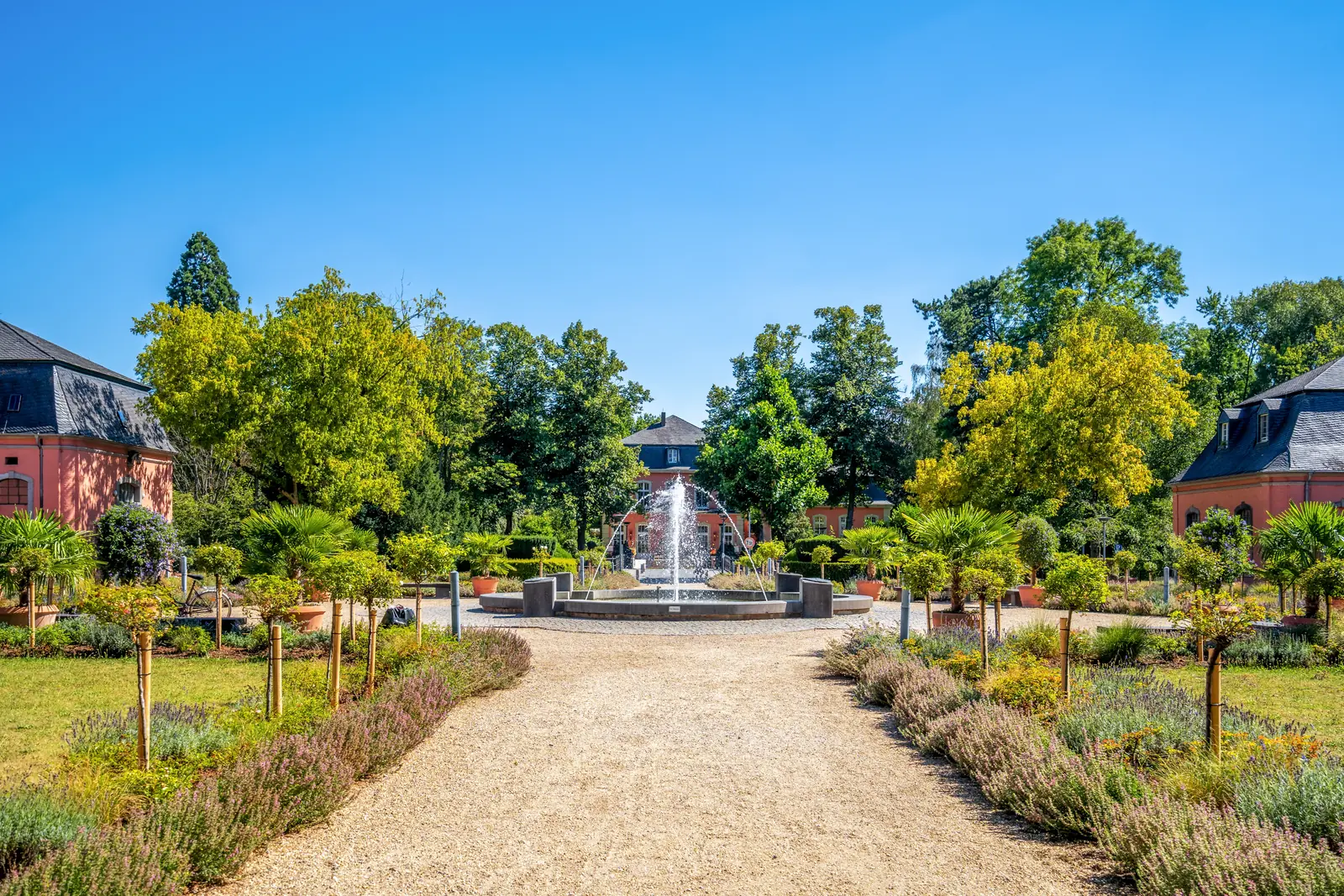 An einem sonnigen Tag führt ein Kiesweg in einem Landschaftsgarten in Mönchengladbach zu einem runden Springbrunnen, der von Bäumen, Sträuchern und rosafarbenen Gebäuden umgeben ist – eine ruhige Kulisse, die sich ideal für besinnliche Gedanken oder sogar für Gespräche über die Betriebsmittelprüfung eignet.