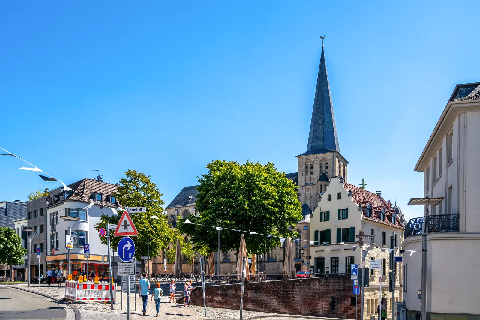 Eine Straßenszene in Mönchengladbach mit Passanten, Verkehrsschildern und einer Kirche mit hohem Kirchturm unter strahlend blauem Himmel vermittelt eine lebendige Stadtatmosphäre. Inmitten des Alltags sorgen regelmäßige Betriebsmittelprüfungen für Sicherheit und Zuverlässigkeit in den Unternehmen der Region.