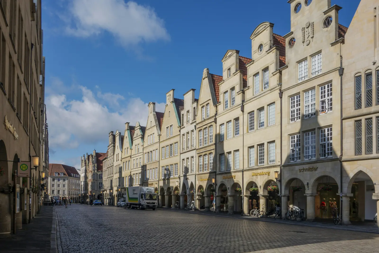 Eine Reihe historischer Giebelhäuser mit gewölbten Durchgängen entlang einer Kopfsteinpflasterstraße unter blauem Himmel in Münster, mit charmanten Ladenfronten, Fahrrädern und einem Lieferwagen – ein Bild, das die lebendige Atmosphäre der Stadt einfängt, in der sich das Alltagsleben und sogar die Betriebsmittelprüfung nahtlos in den urbanen Rhythmus einfügen.