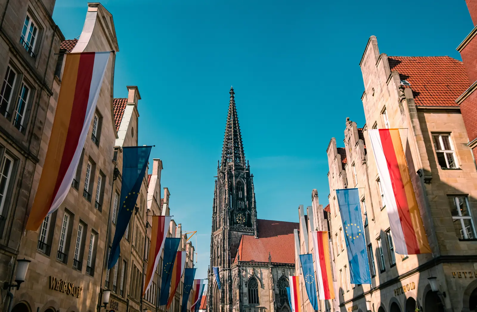 Eine gotische Kirche mit einem hohen Turm, die inmitten historischer Gebäude in Münster steht, die alle mit verschiedenen europäischen Landesflaggen geschmückt sind, unter einem strahlend blauen Himmel.