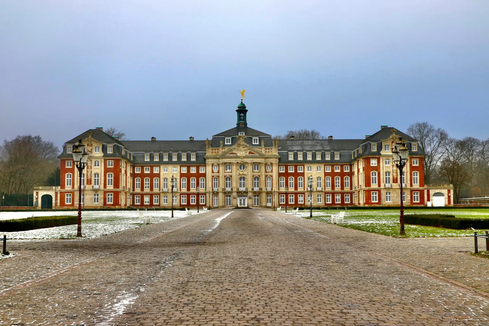 Ein Schloss im Barockstil mit einem zentralen Turm, einer rot-beigen Fassade, einem Kopfsteinpflasterweg und einer dünnen Schneeschicht auf dem Boden, gelegen in der Nähe von Münster und umgeben von Bäumen unter einem bewölkten Himmel – eine malerische Kulisse, in der moderne Anforderungen wie die Betriebsmittelprüfung auf historischen Charme treffen.
