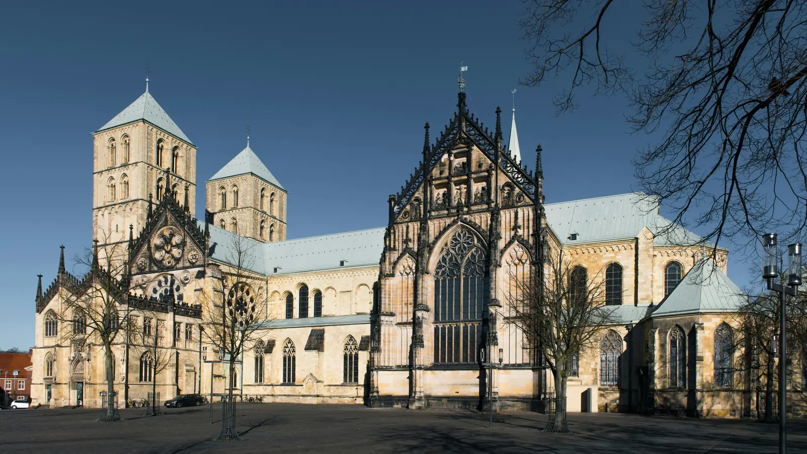 Eine große Steinkathedrale, die an das beeindruckende Münster erinnert, mit zahlreichen Türmen, Bogenfenstern und detailreichen architektonischen Elementen im gotischen und romanischen Stil, vor dem Hintergrund eines strahlend blauen Himmels.