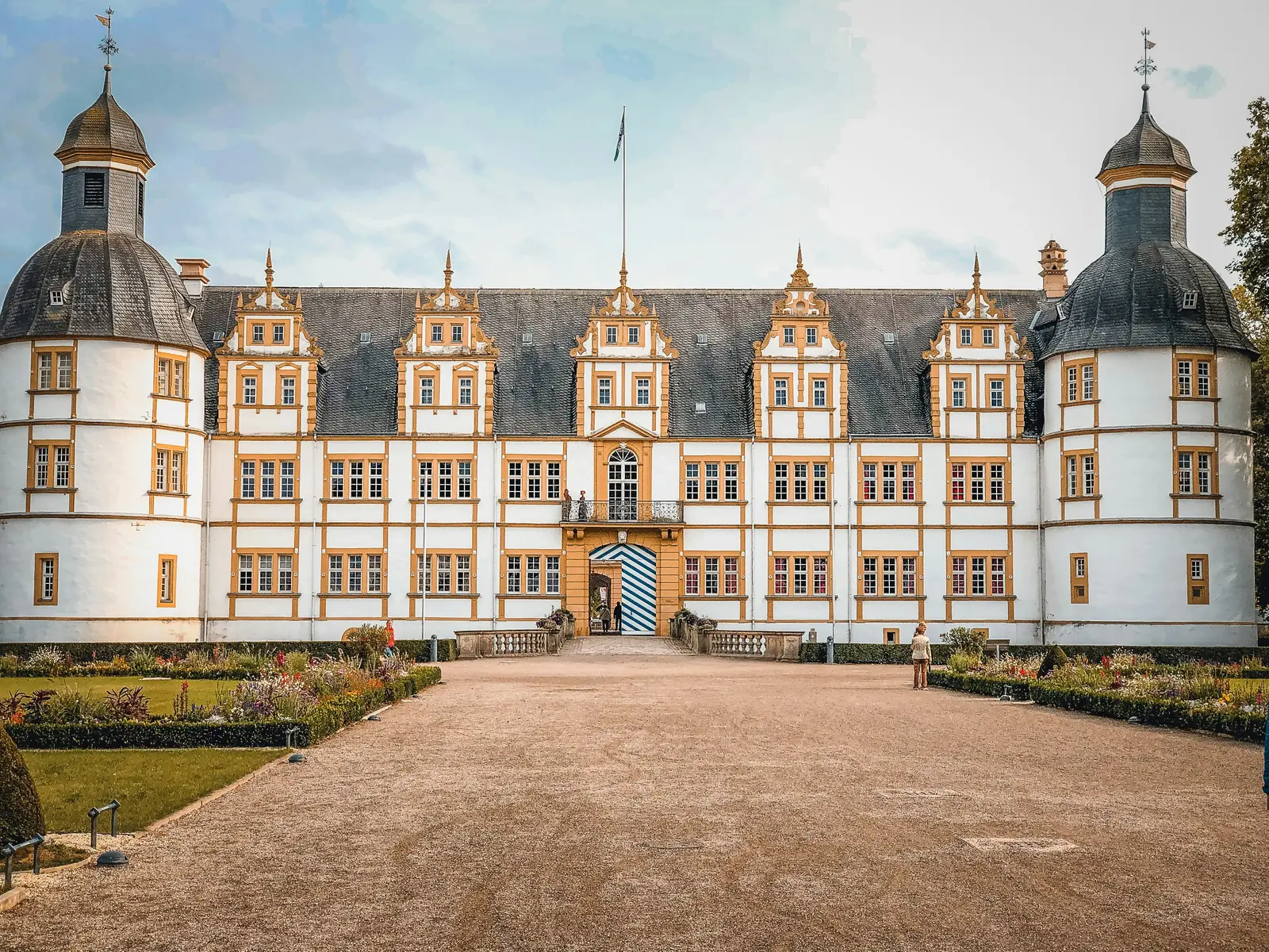Ein großes, symmetrisches historisches Gebäude mit weißen Wänden, gelben Zierleisten und zwei runden Türmen steht hinter einem Garten und einem Kiesweg unter einem teilweise bewölkten Himmel in Münster und vermittelt eine Atmosphäre, in der Tradition auf moderne Anforderungen wie die Betriebsmittelprüfung trifft.