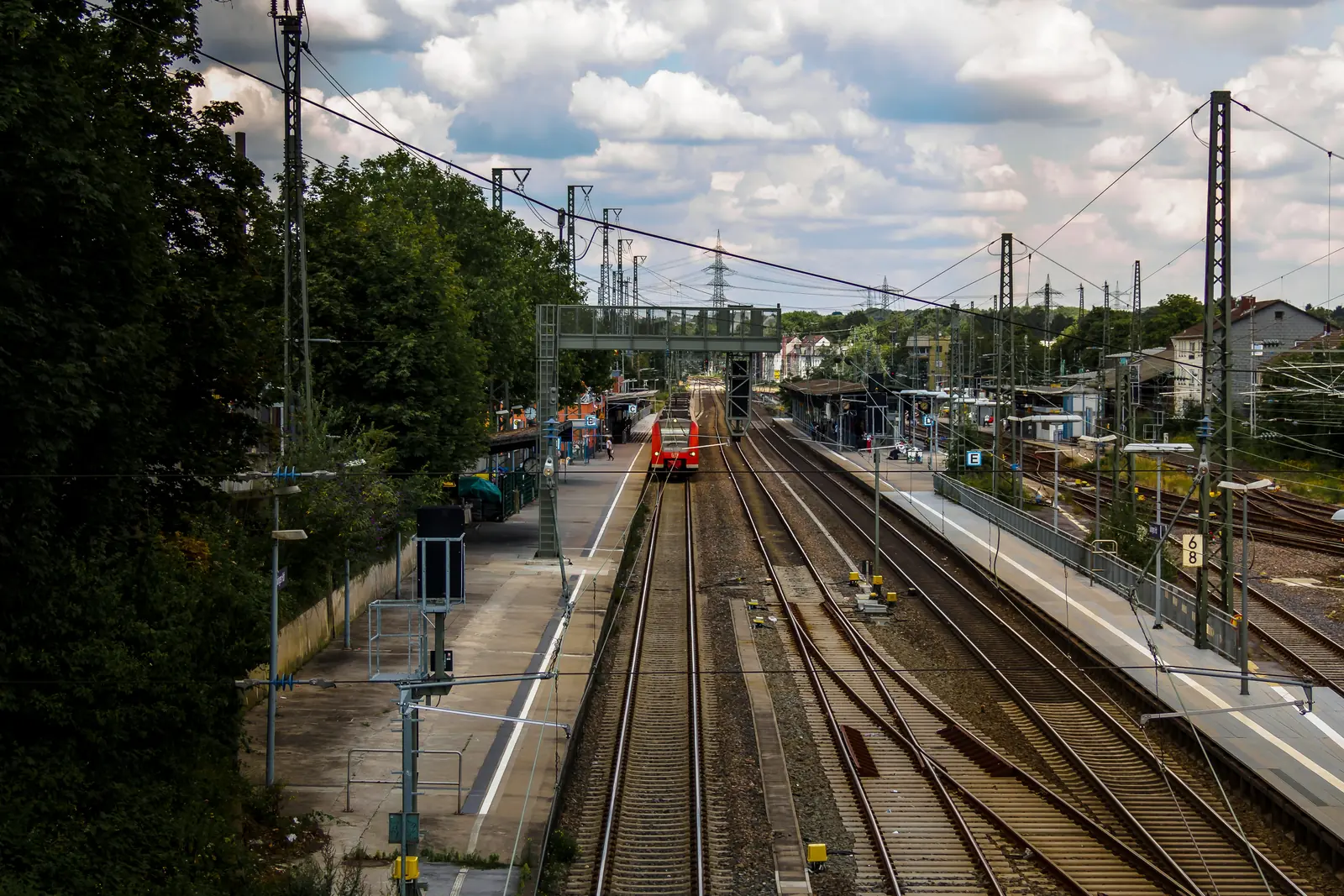 Ein roter Zug nähert sich einem Bahnhof in Solingen mit mehreren Gleisen und Bahnsteigen, umgeben von Bäumen, Oberleitungen und bewölktem Himmel.