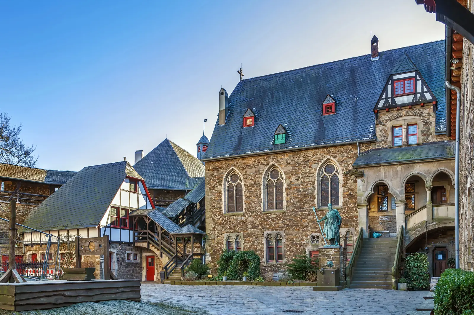 Ein historisches Steingebäude mit Bogenfenstern, einem steilen Schieferdach und einer zentralen Treppe steht in einem mit Kopfsteinpflaster gepflasterten Innenhof, in dessen Nähe sich eine Statue und Fachwerkhäuser befinden, die den traditionellen Charme Solingens widerspiegeln.