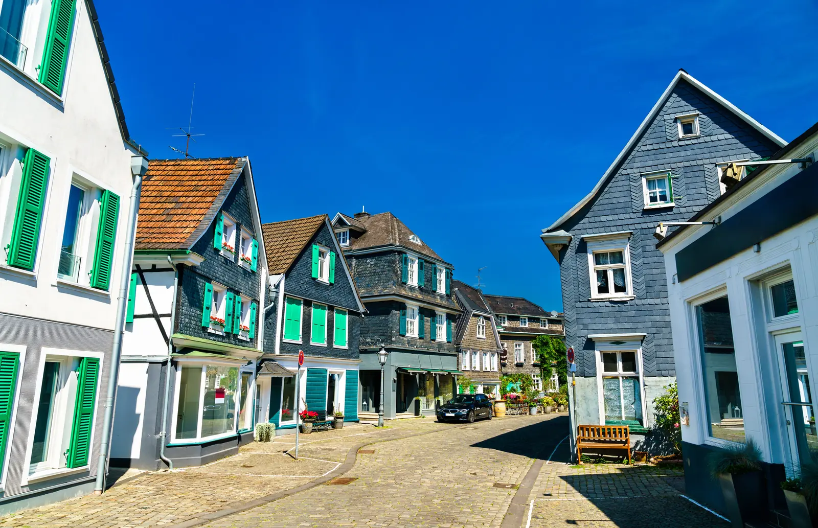 Eine ruhige Kopfsteinpflasterstraße in Solingen, gesäumt von traditionellen europäischen Häusern mit grünen Fensterläden und Satteldächern unter strahlend blauem Himmel, bietet ein malerisches Bild, das im Kontrast zur industriellen Seite der Stadt steht, die für ihre Betriebsmittelprüfung bekannt ist.