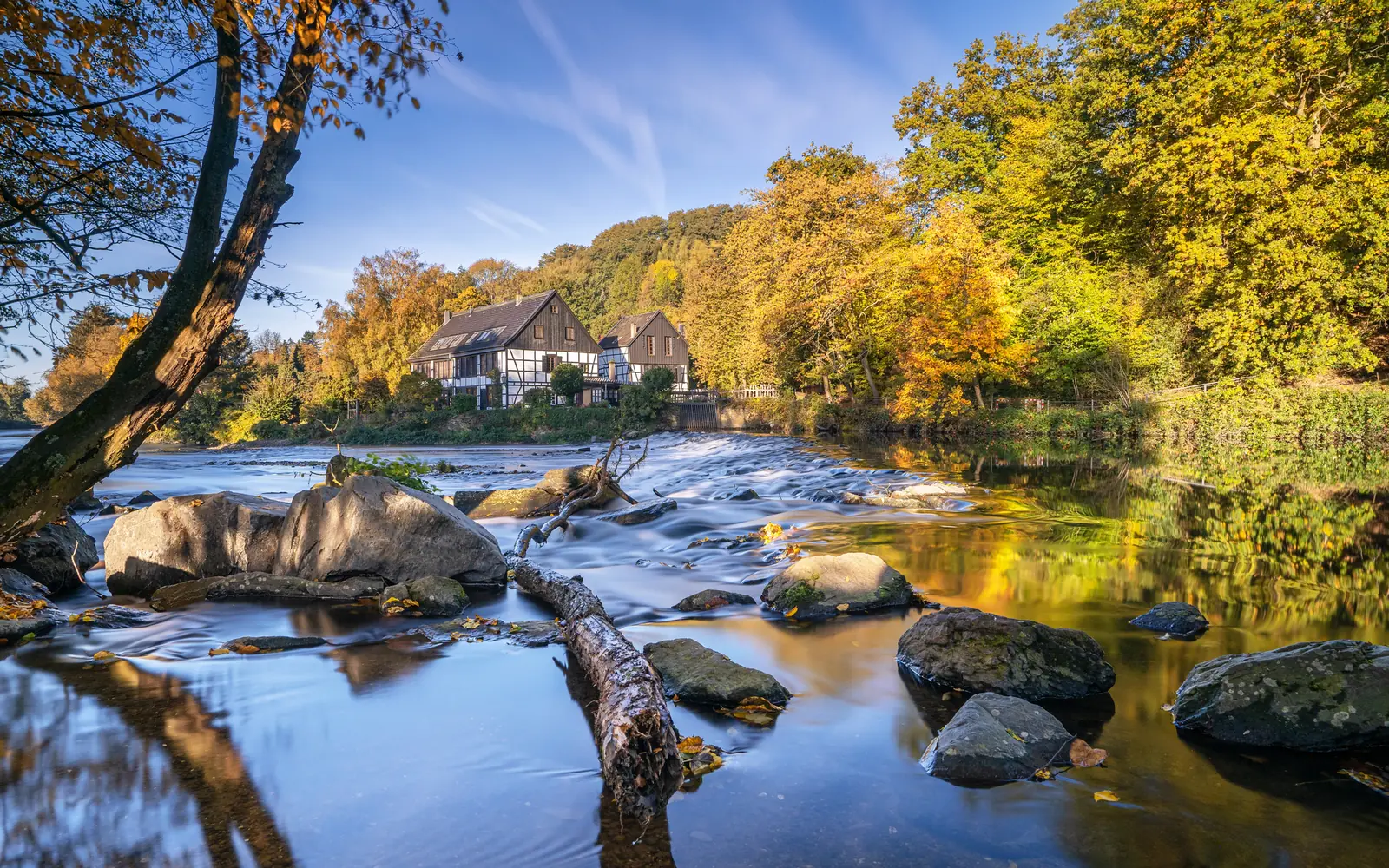 In Solingen steht ein Steinhaus an einem ruhigen Fluss mit Felsen und einem sanften Wasserfall, umgeben von Bäumen mit herbstlichem Laub unter einem strahlend blauen Himmel.
