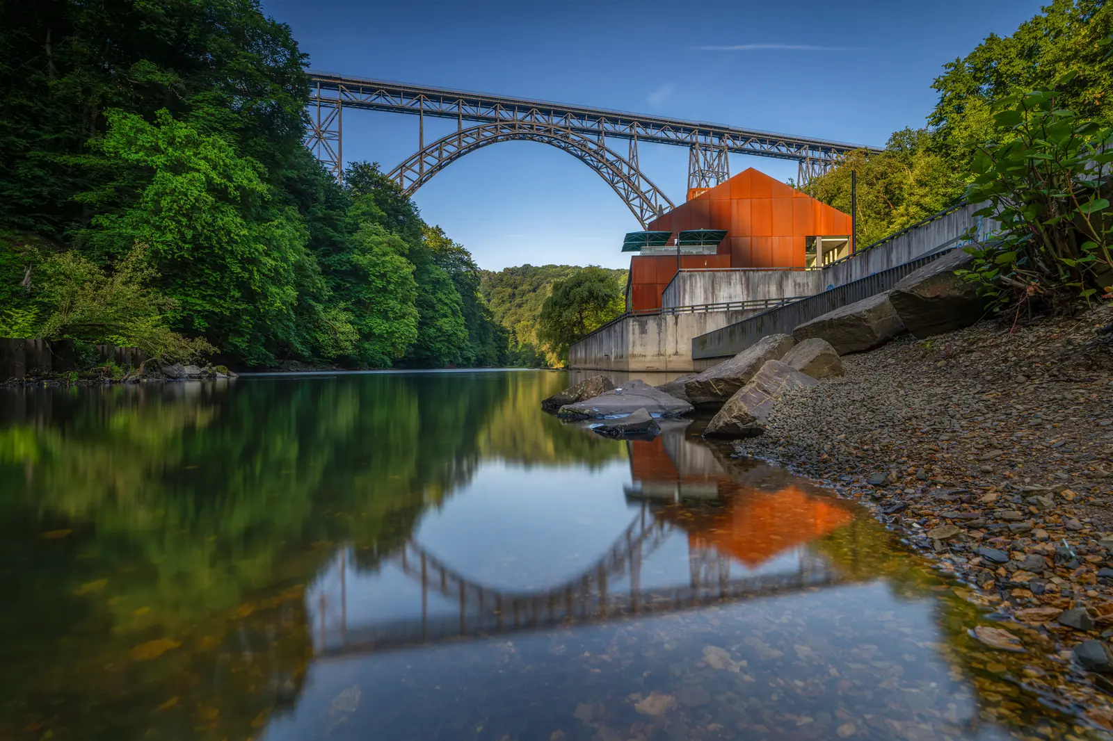 Eine Stahlbogenbrücke überspannt einen Fluss, der von dichtem Grün umgeben ist; im Vordergrund sind ein modernes orangefarbenes Gebäude und ein felsiges Ufer zu sehen, die sich alle wunderschön im ruhigen Wasser spiegeln – eine malerische Szene, die an Solingen erinnert.