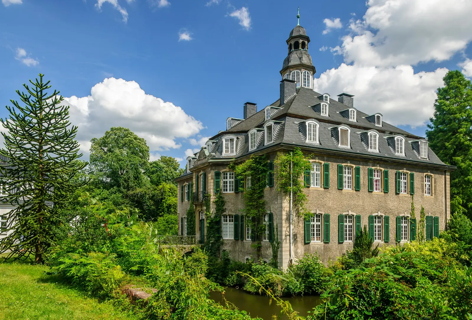 Ein historisches Herrenhaus im europäischen Stil mit grünen Fensterläden, Kletterpflanzen und einer markanten Kuppel – das an die eleganten Anwesen in Solingen erinnert –, umgeben von üppiger Vegetation unter einem teilweise bewölkten Himmel.
