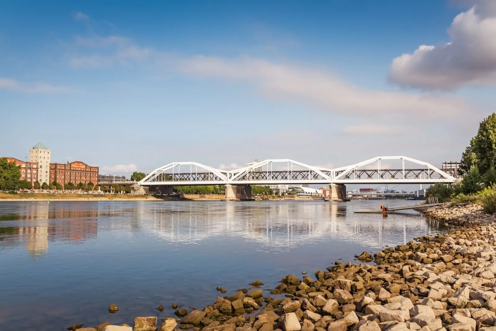 Weiße Bogenbrücke über den Rhein bei Ludwigshafen mit Steinufer im Vordergrund.