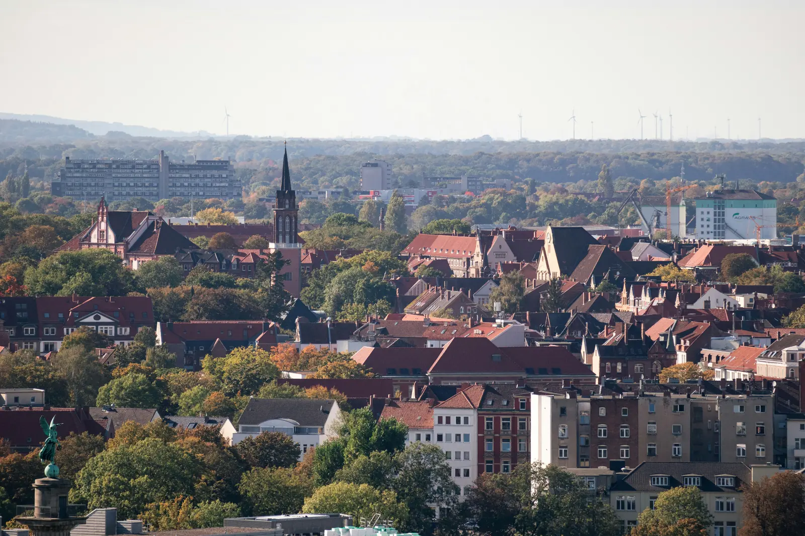 Blick über die Dächer von Hannover mit einem schlanken Kirchturm und Windrädern am Horizont.