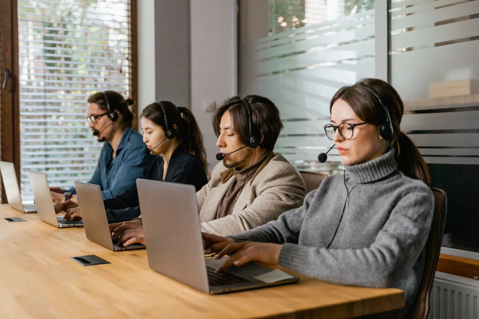 Betriebsmittelprüfung im Callcenter: Mitarbeitende mit Headsets und Laptops am langen Tisch.