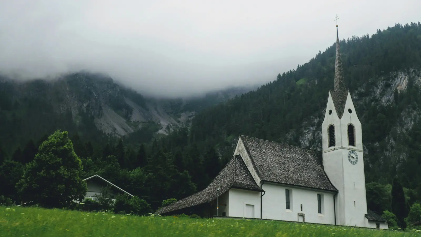 Betriebsmittelprüfung und Leiter- und Trittprüfung in der Kirche: Kirche mit spitzem Turm vor bewaldeten Hängen.