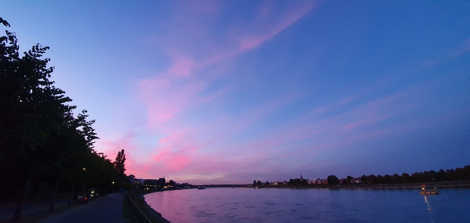 Ein Fluss in der Abenddämmerung mit rosa und blauen Wolken am Himmel, Bäumen am linken Ufer und Gebäuden, die in der Ferne auf der rechten Seite zu sehen sind.