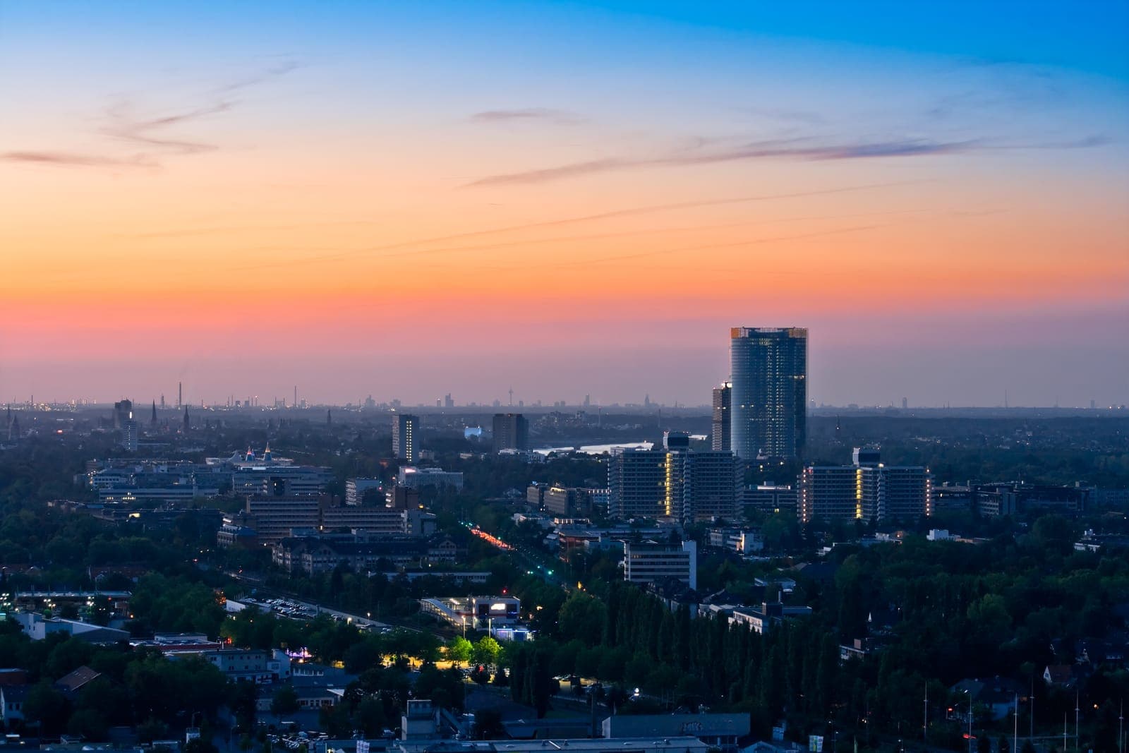 Stadtansicht bei Sonnenuntergang mit einem hohen Gebäude und mehreren mittelhohen Gebäuden, Bäumen im Vordergrund und einem orange-blauen Farbverlauf am Himmel im Hintergrund.