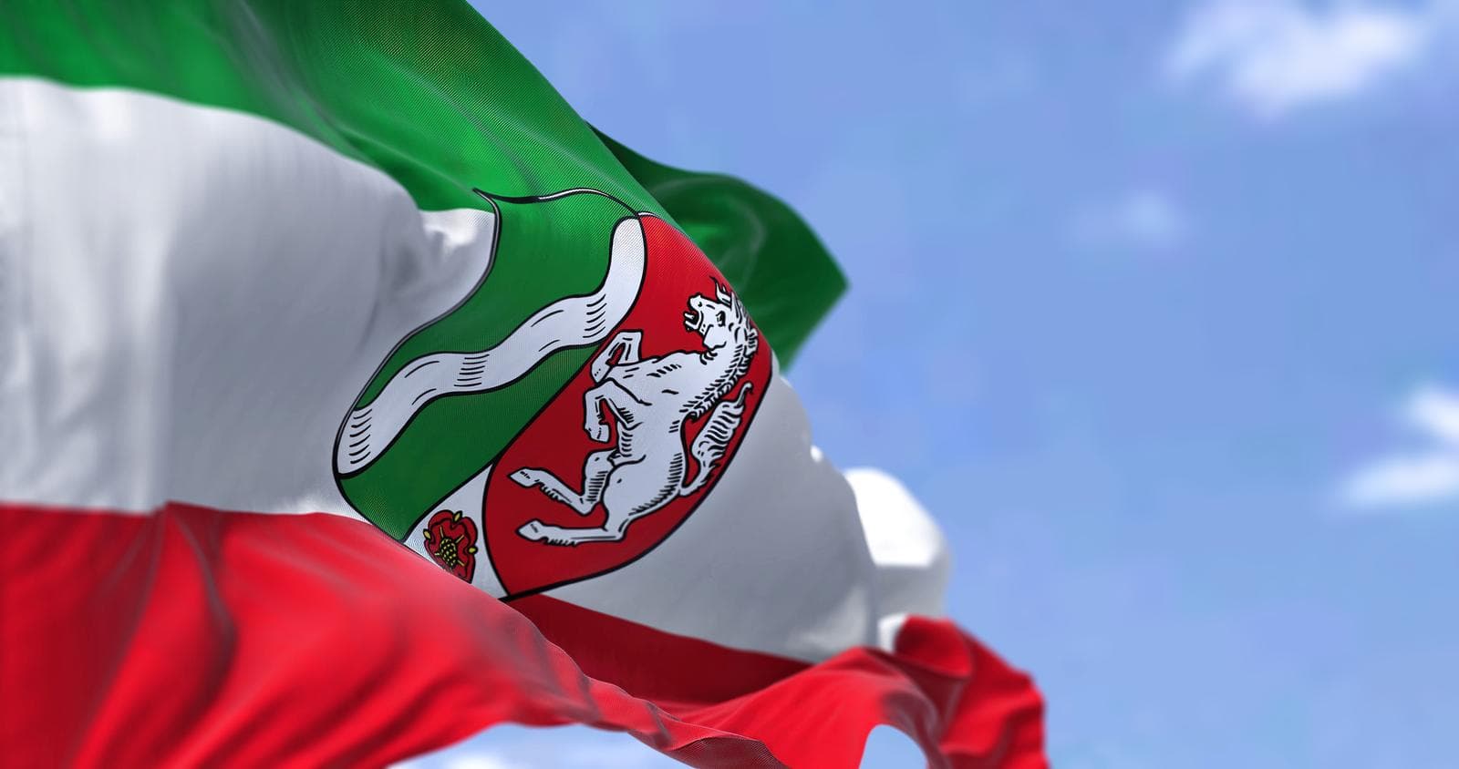 Die Flagge von Nordrhein-Westfalen, Deutschland, mit horizontalen grünen, weißen und roten Streifen sowie einem Wappen, die vor einem blauen Himmel weht.