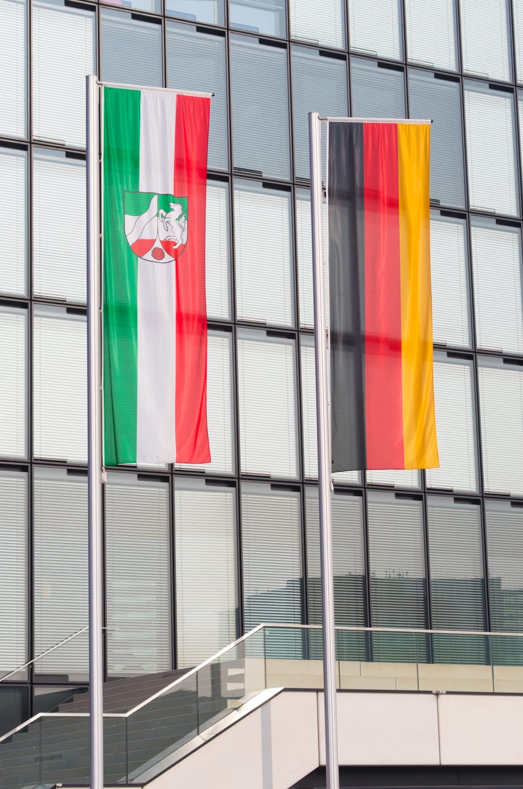 Zwei Flaggen an Fahnenmasten vor einem modernen Glasgebäude: die Flagge von Nordrhein-Westfalen (links) und die deutsche Nationalflagge (rechts).