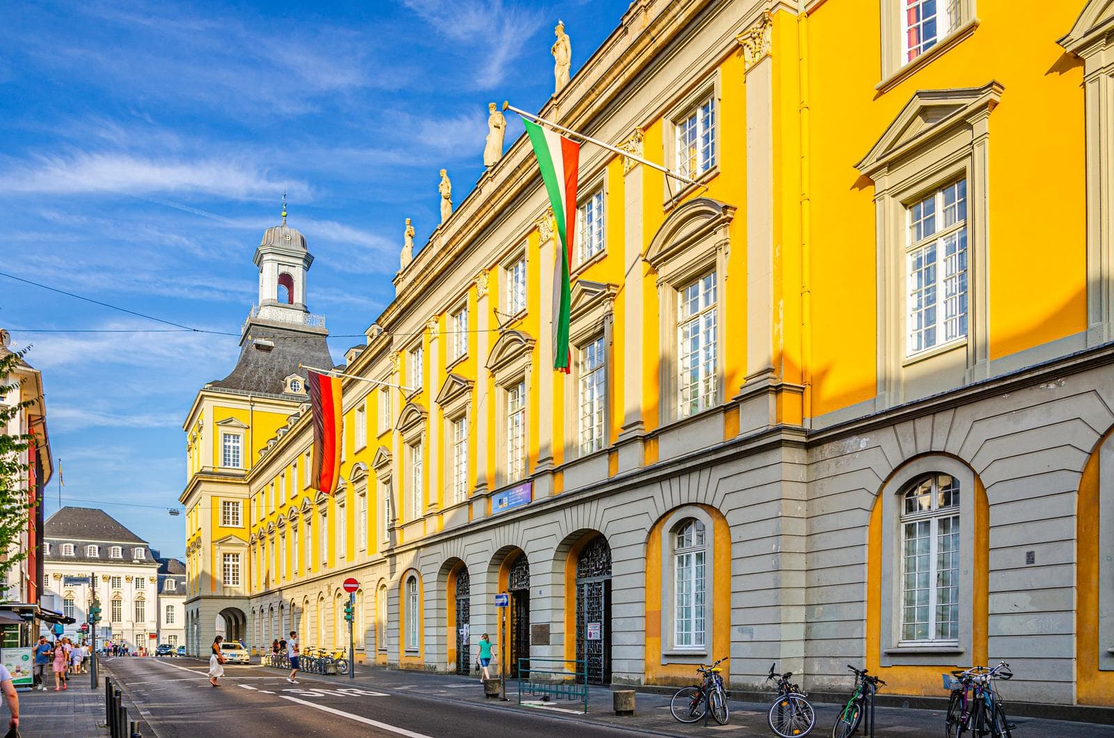 Ein großes, gelbes, historisches Gebäude mit Bogenfenstern und Fahnen, gelegen an einer Straße mit geparkten Fahrrädern unter strahlend blauem Himmel.
