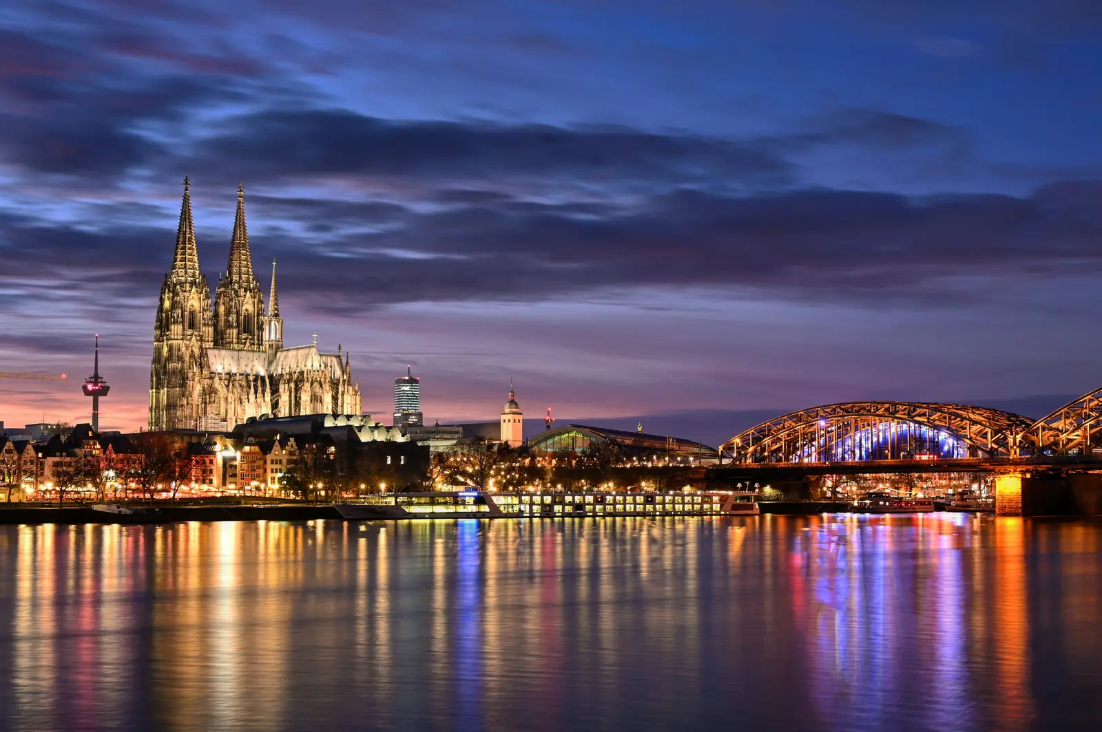 Kölner Dom und Hohenzollernbrücke spiegeln sich abends im Rhein.